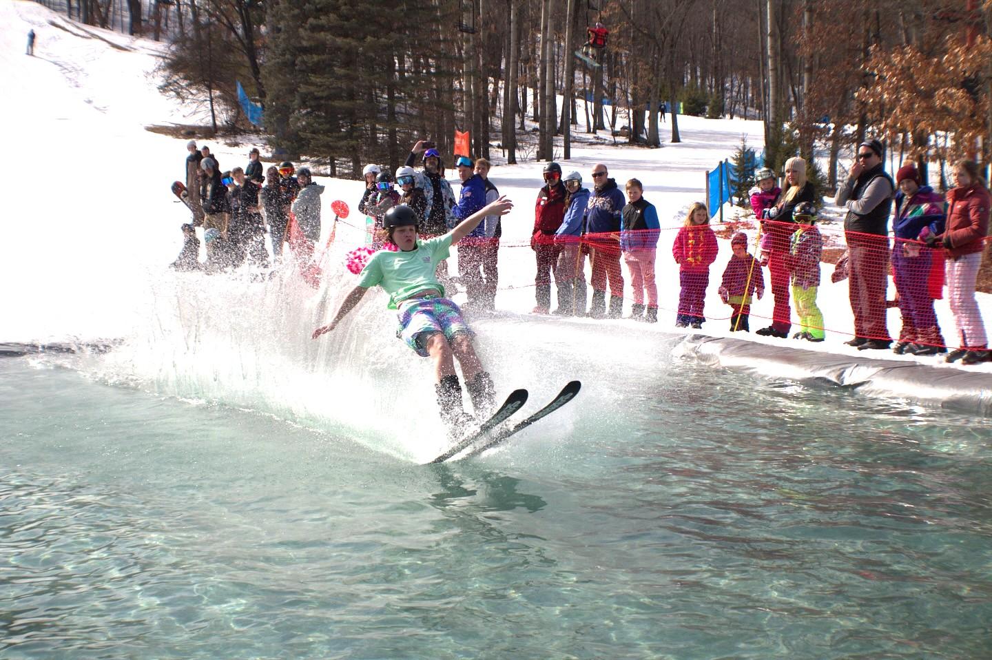 Skier skims across a water pool, surrounded by spectators in winter clothing.