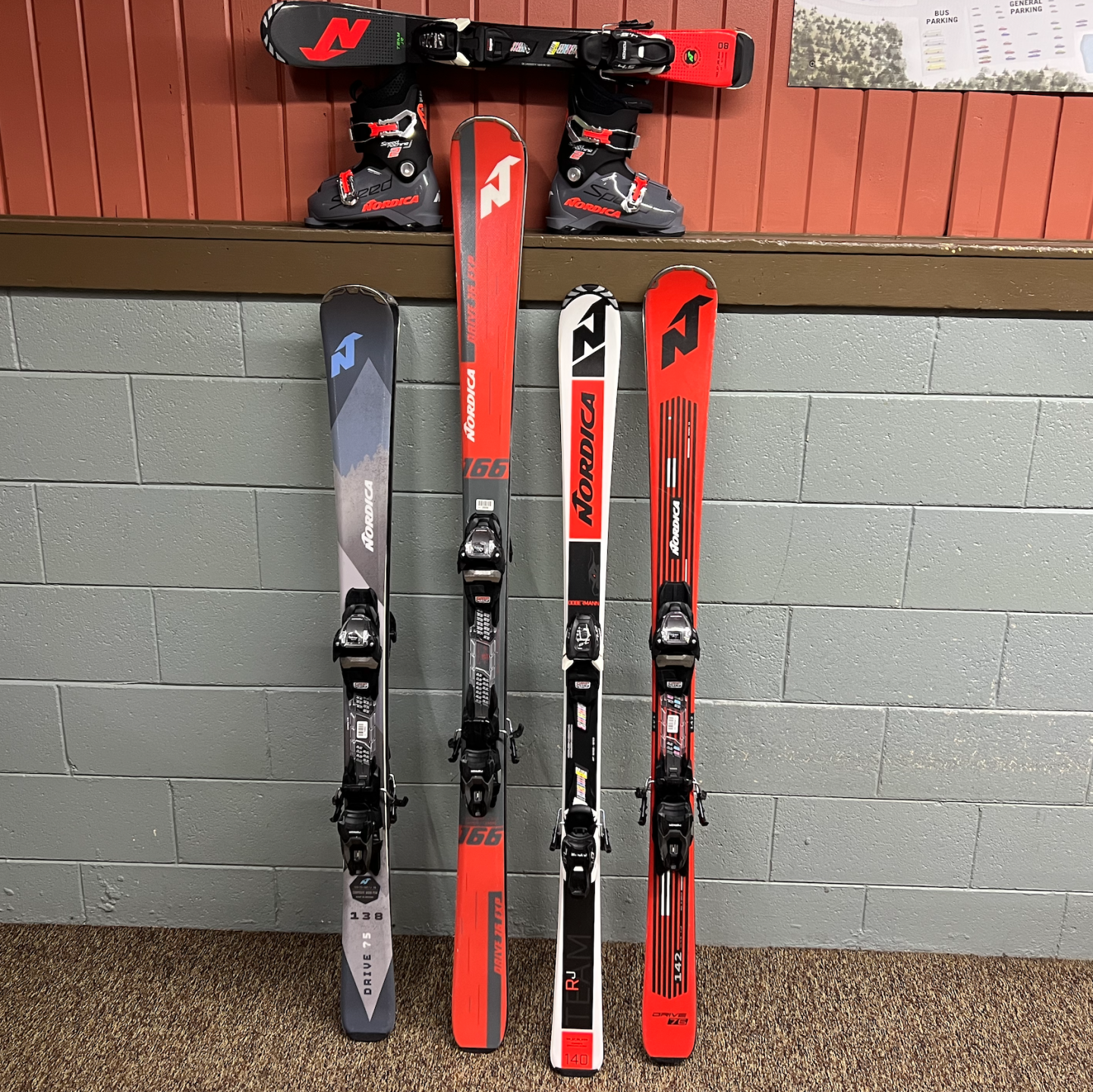 Three pairs of skis and two ski boots against a wall.