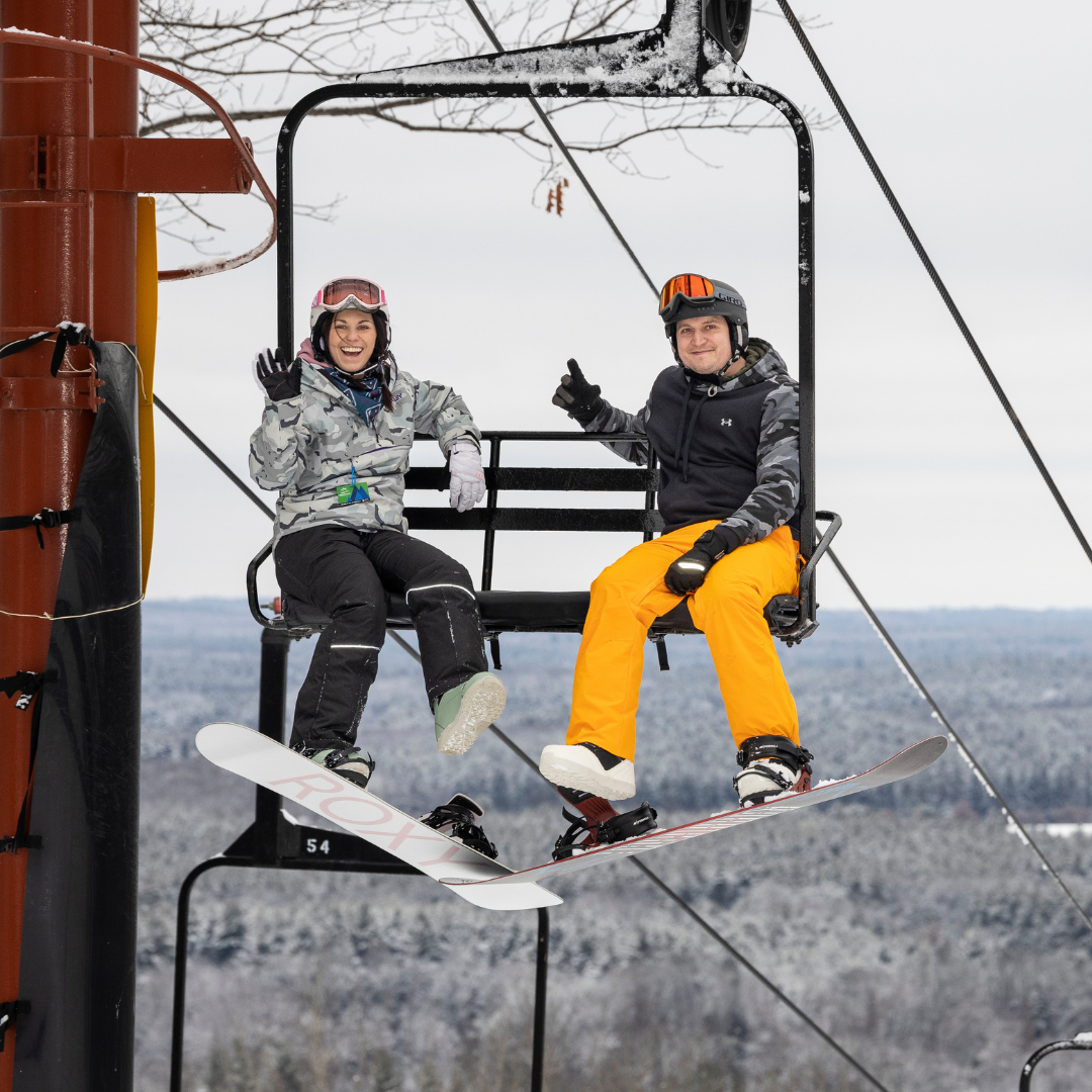 Two people on a ski lift, wearing snow gear, smiling and waving.