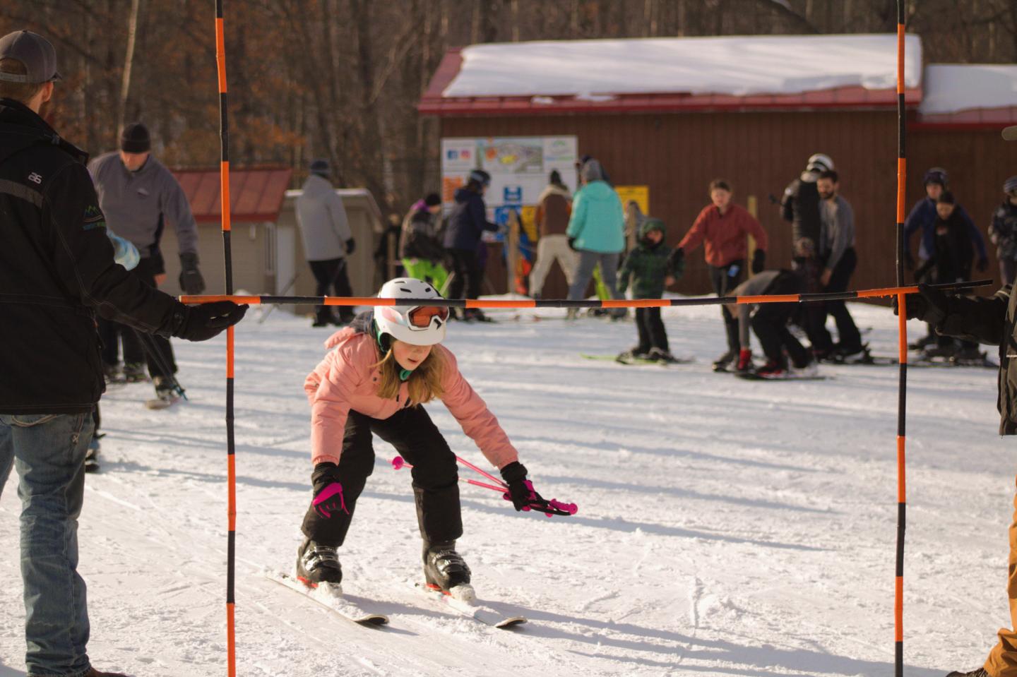 Child skiing under orange poles, surrounded by people on a snowy day.