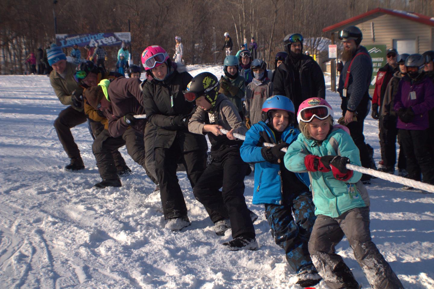 A group of people playing tug-of-war on snow in winter gear.