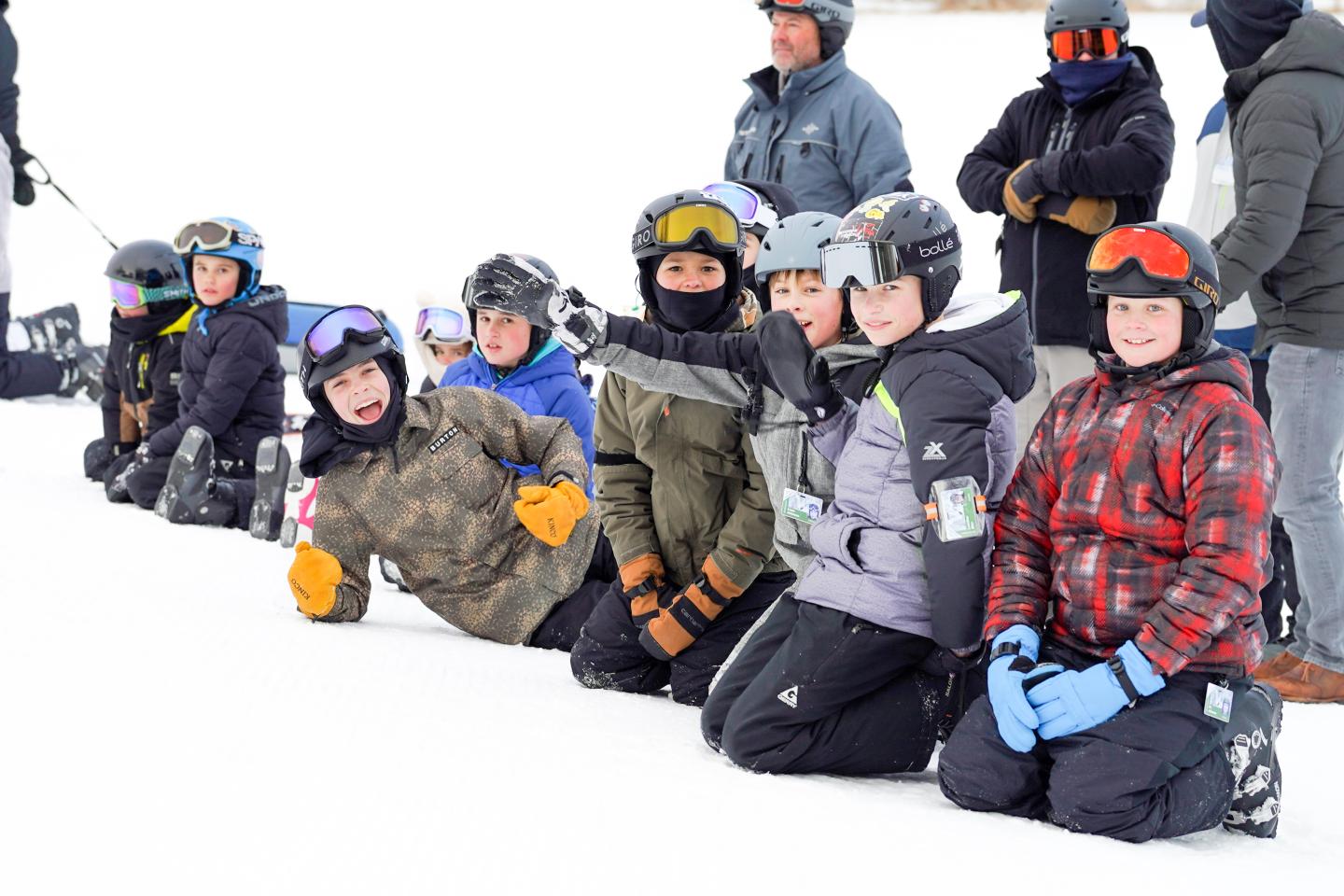 Children in winter gear kneeling in snow, smiling and wearing helmets.