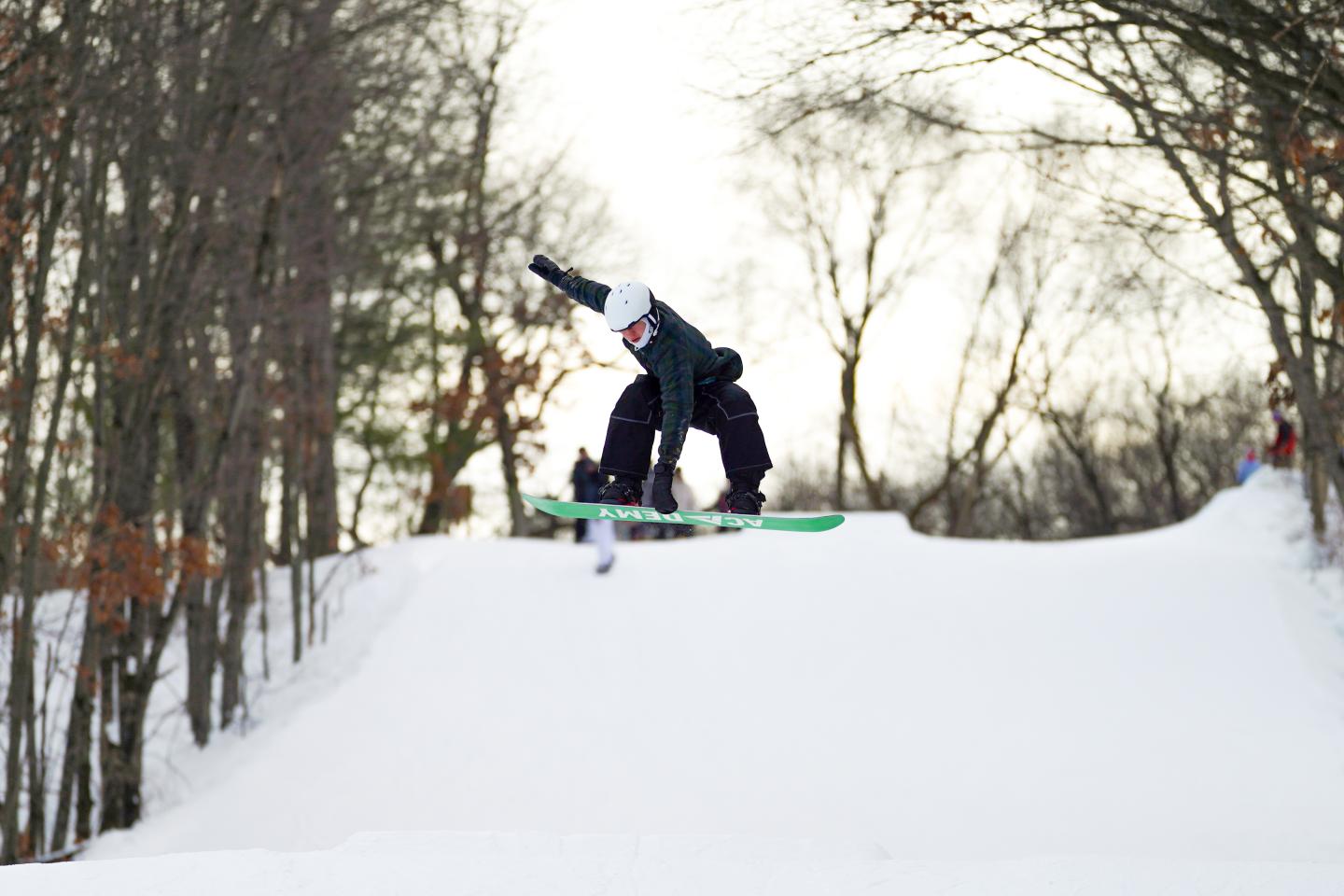 Snowboarder mid-air on a snowy slope next to leafless trees.