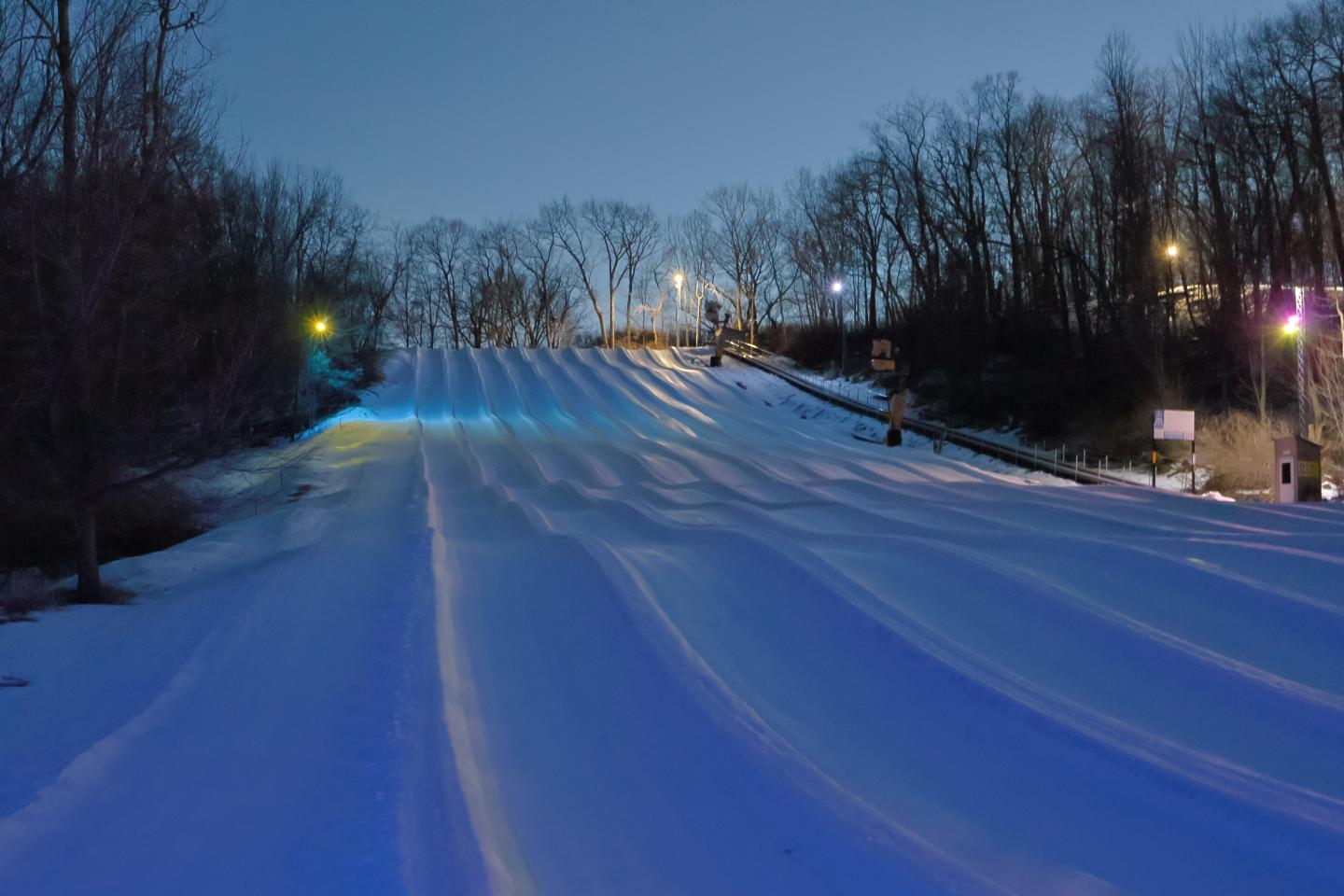Snow-covered hill at night, lit with blue and yellow lights, surrounded by trees.