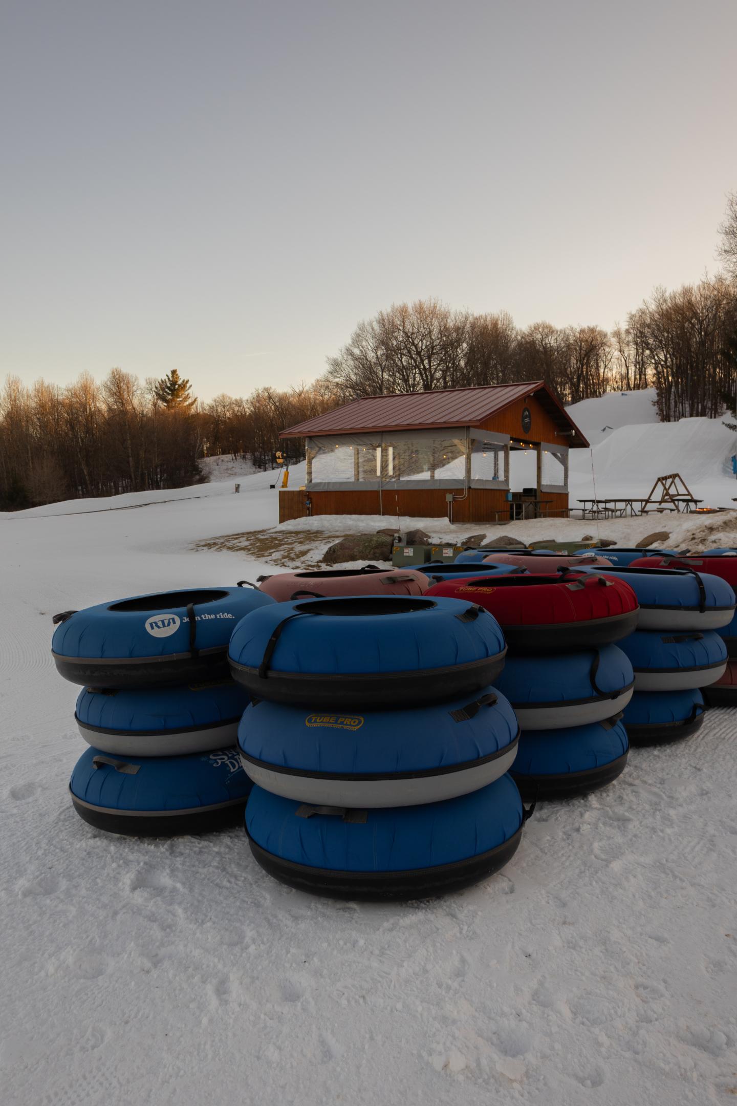 Snow tubes stacked on snowy ground near a cabin at sunset.