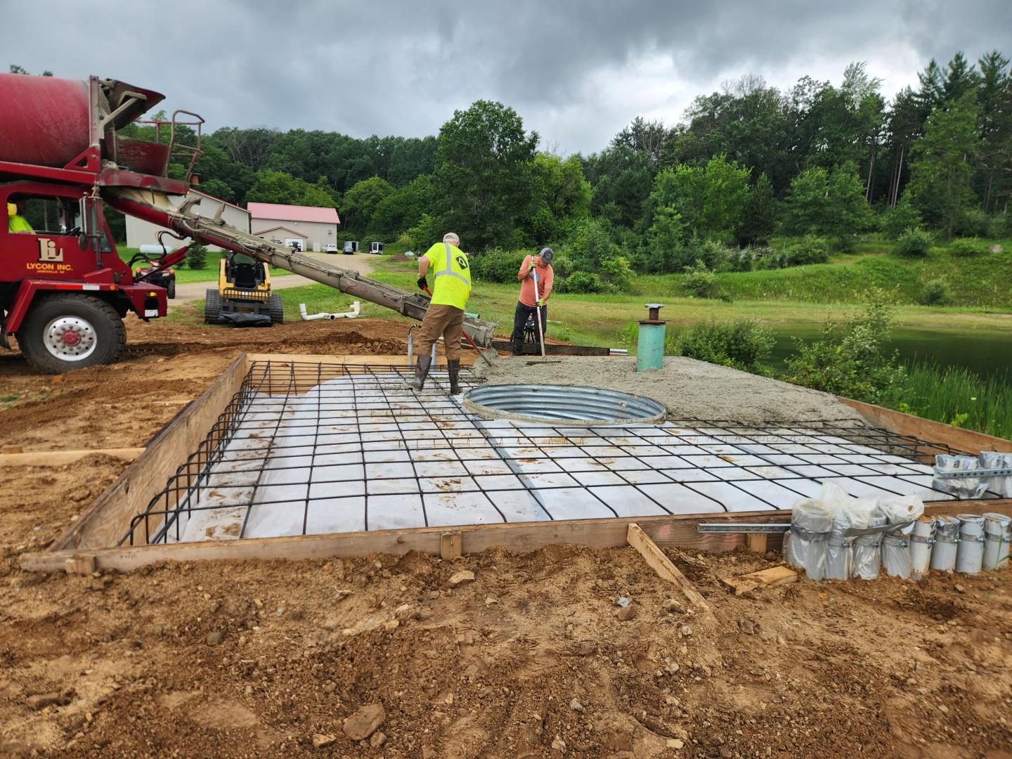 Workers pour concrete over rebar foundation beside a pond under cloudy skies.