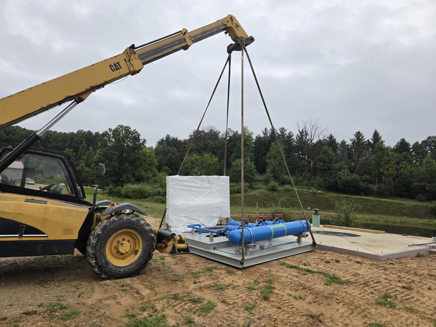 Crane lifting a white wrapped pallet beside construction materials under cloudy skies.