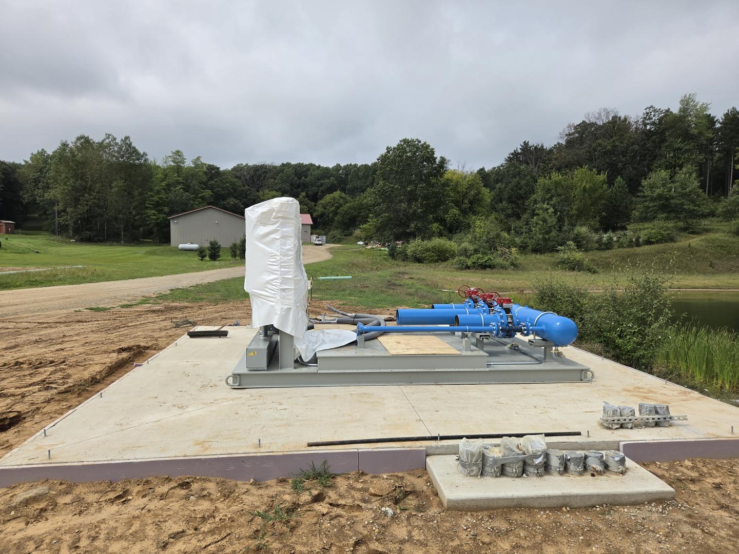 Concrete equipment pad with wrapped machinery and blue pipes at a construction site.