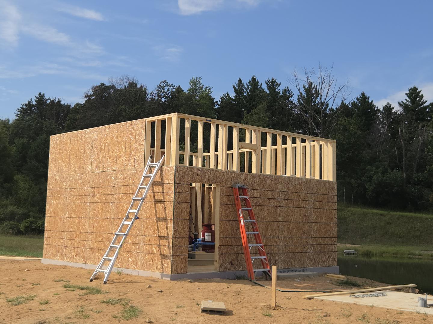 Partially built wooden house frame with ladders on a sandy construction site.