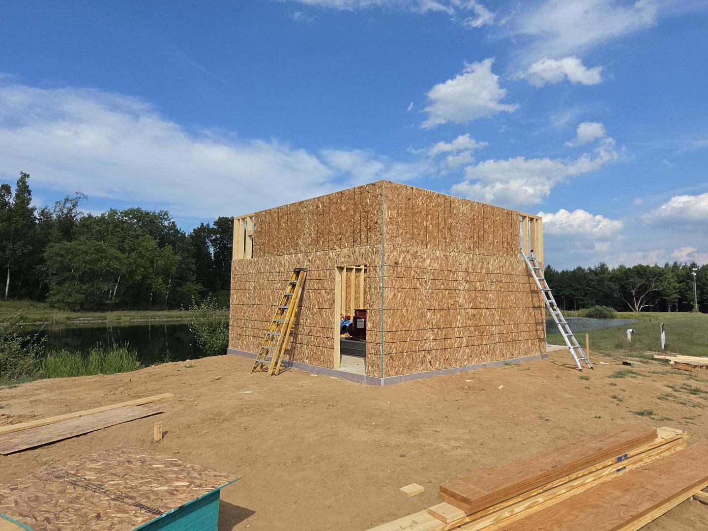 Wood-framed house under construction on a dirt lot beneath a blue sky.