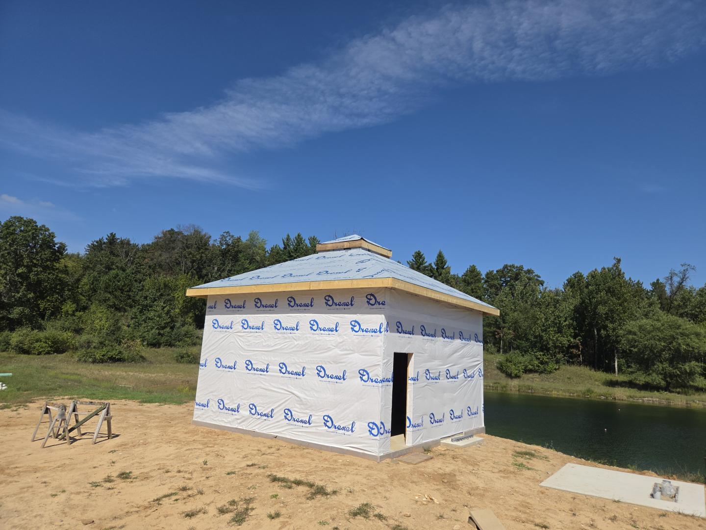 Small house under construction beside a pond, wrapped in blue-logo insulation.
