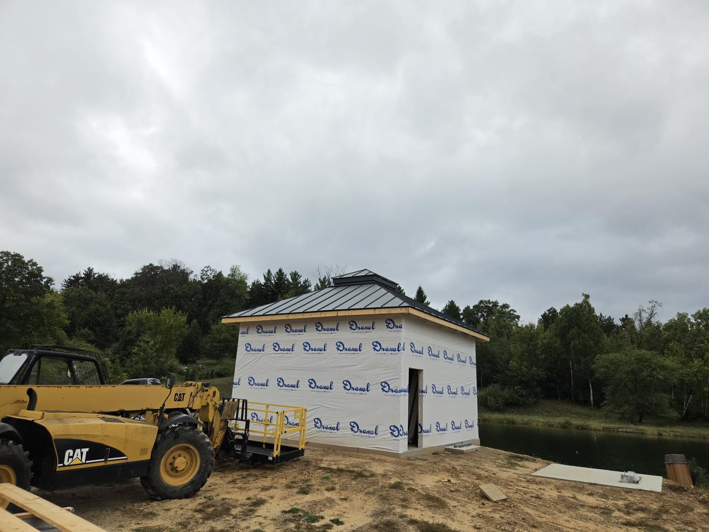 House under construction wrapped in weather barrier, with excavator under cloudy sky.