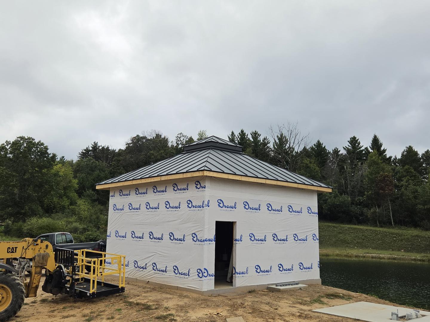 Small white construction building with blue wrap, a black door, and equipment under cloudy skies.