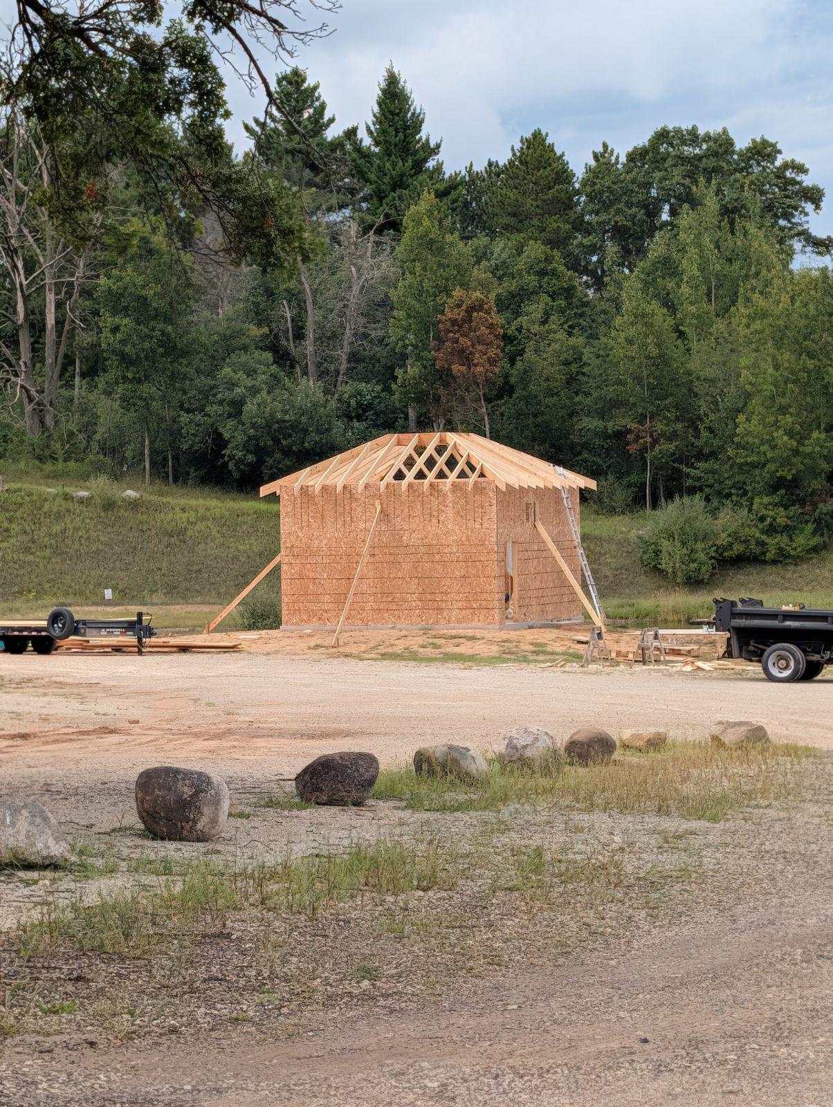 Wood-framed cabin under construction beside parked trailers in a forest clearing.