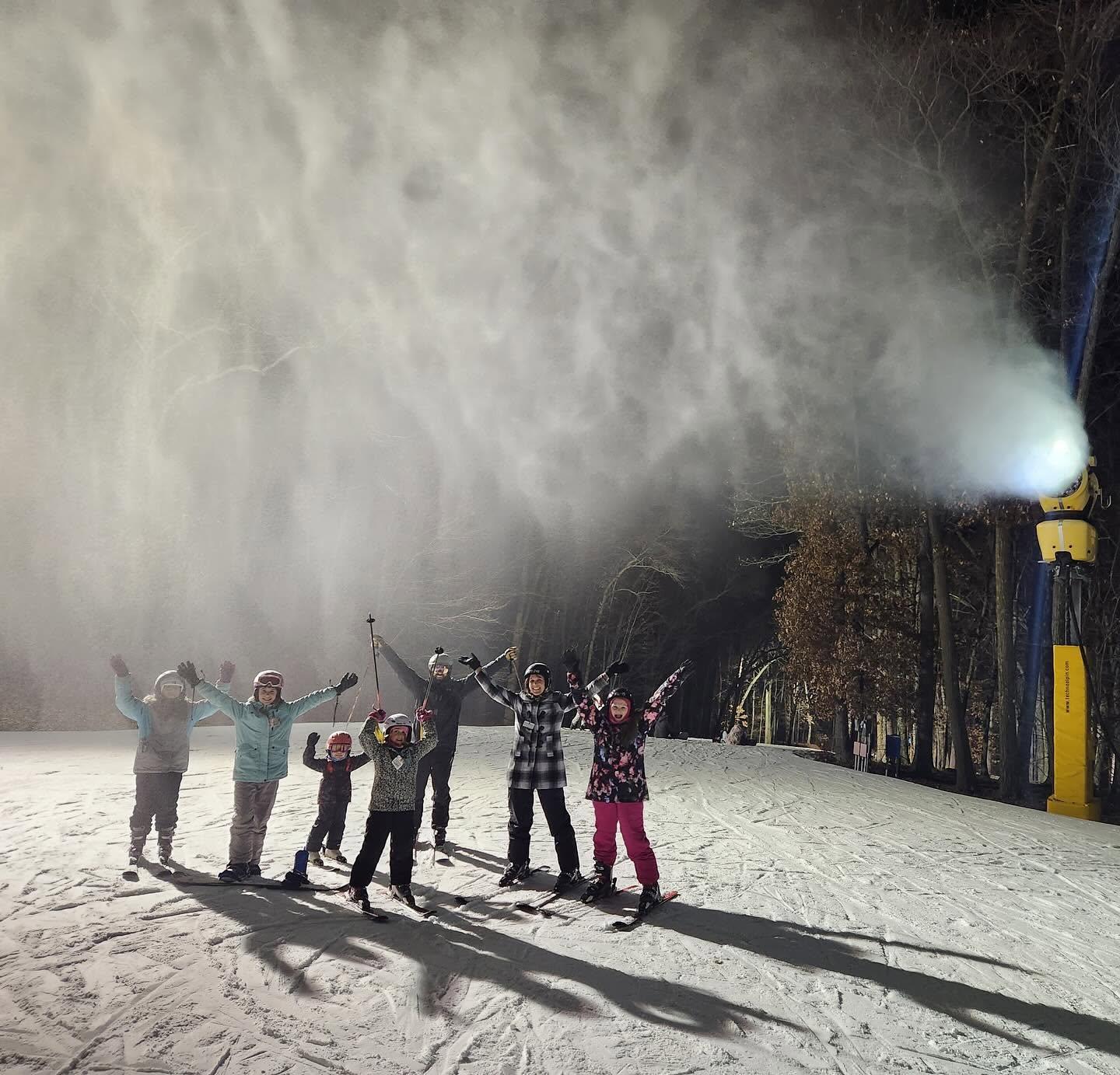 Five snowboarders celebrate on a snowy slope under bright floodlights.