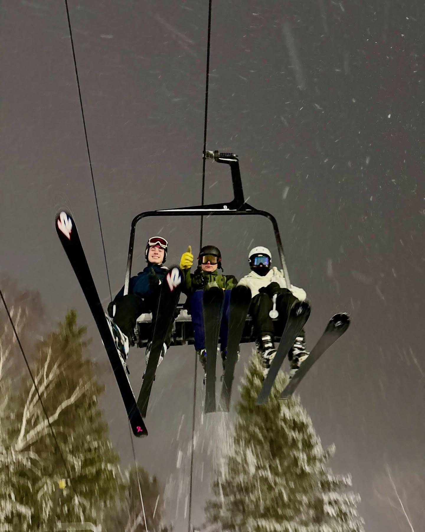 Four skiers riding a chairlift above snowy evergreen trees.
