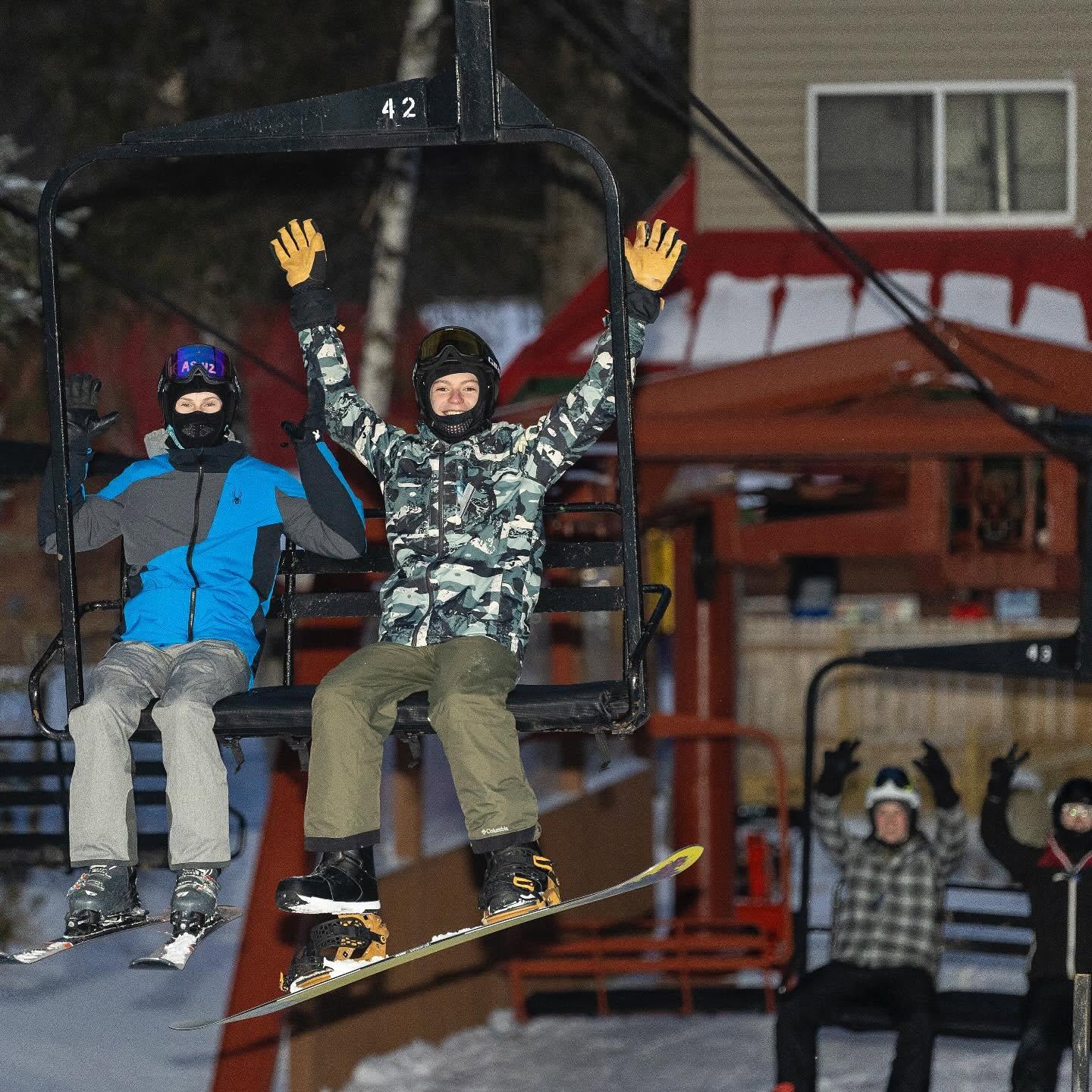 Snowboarders jumping near a ski lift, with one raising both hands.