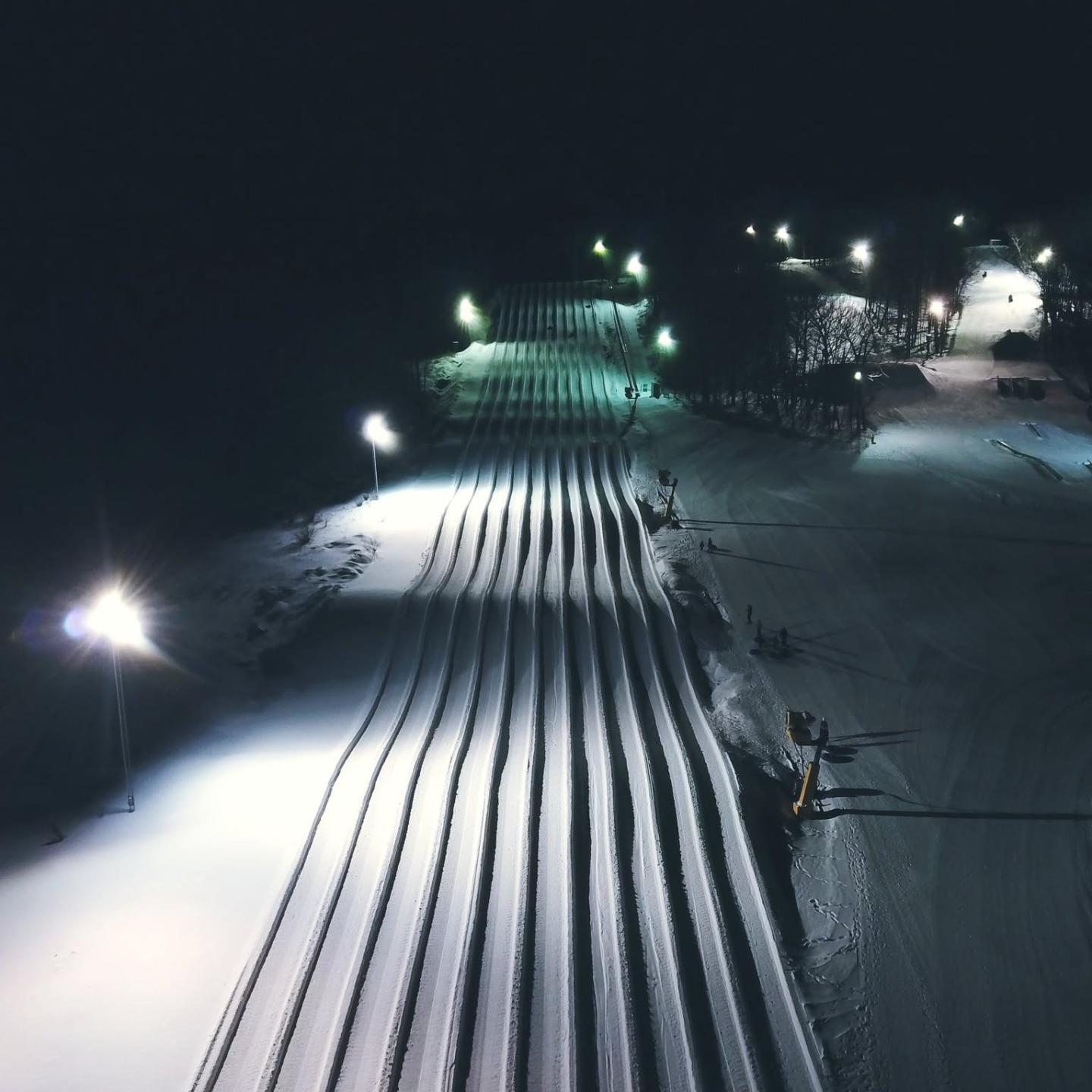 Floodlit ski slope at night with freshly groomed parallel tracks.