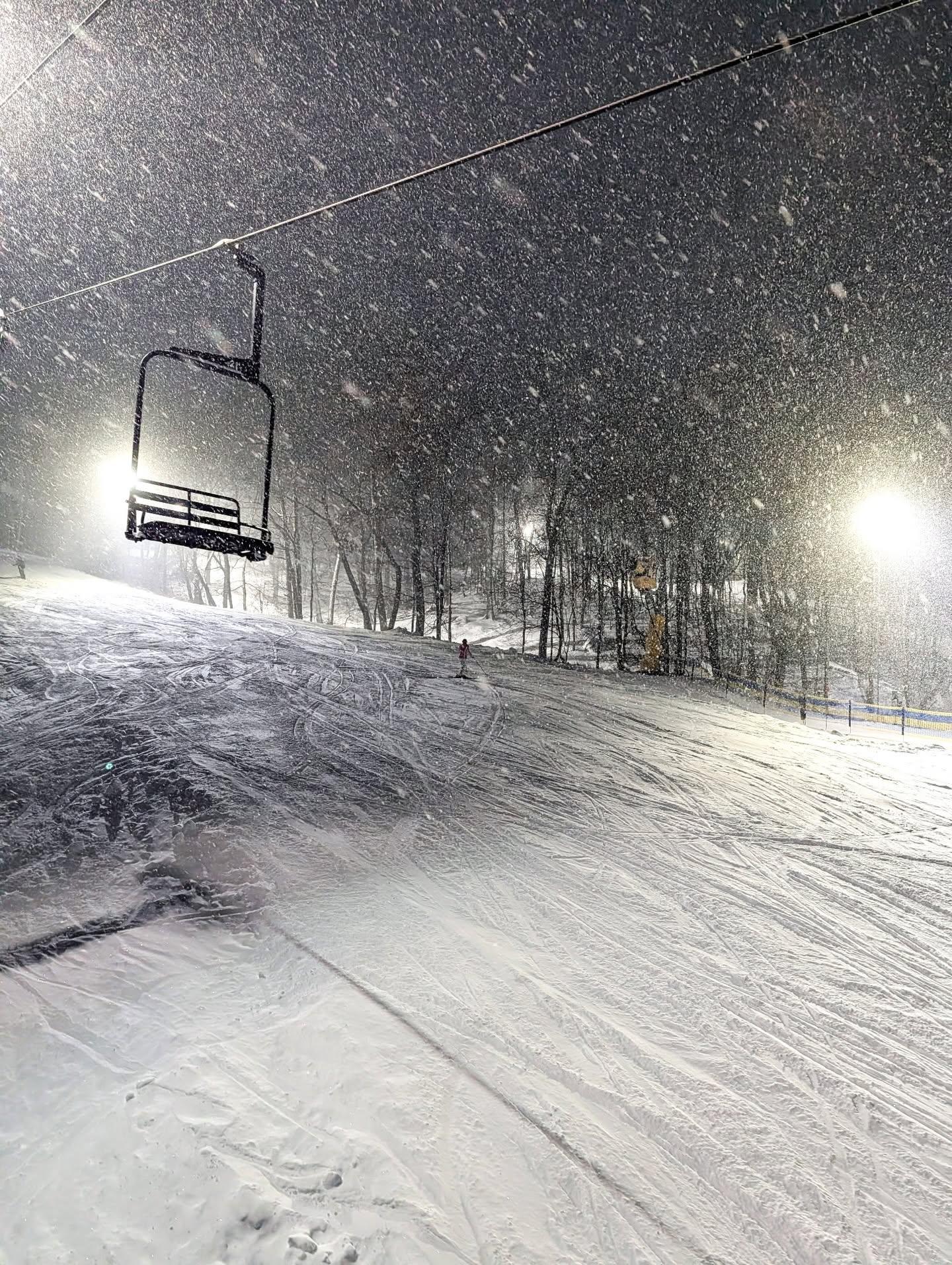 Snowy ski slope at night with an empty chairlift and glowing lights.