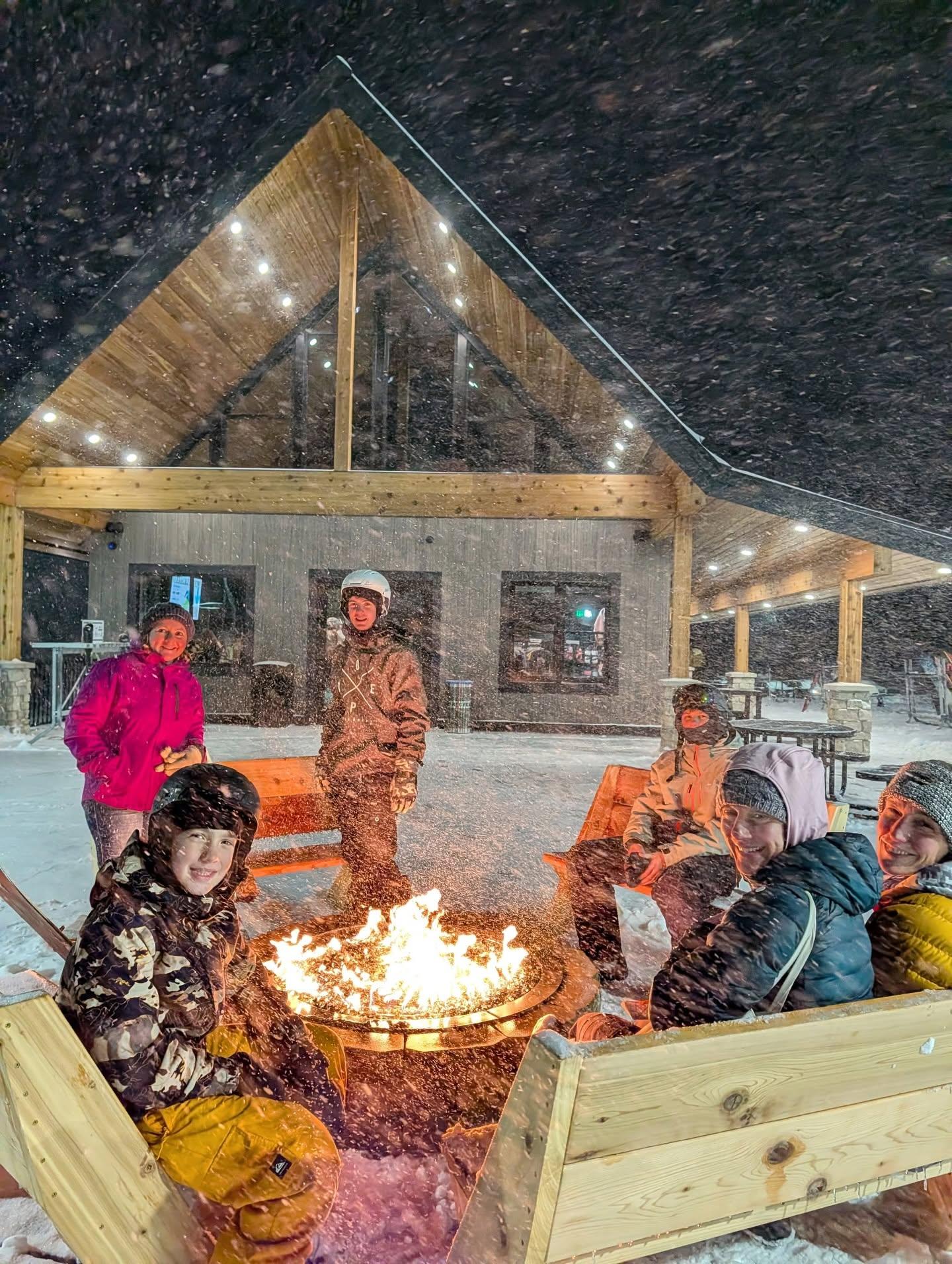 Snowy outdoor gathering around a fire pit beside a lit cabin.