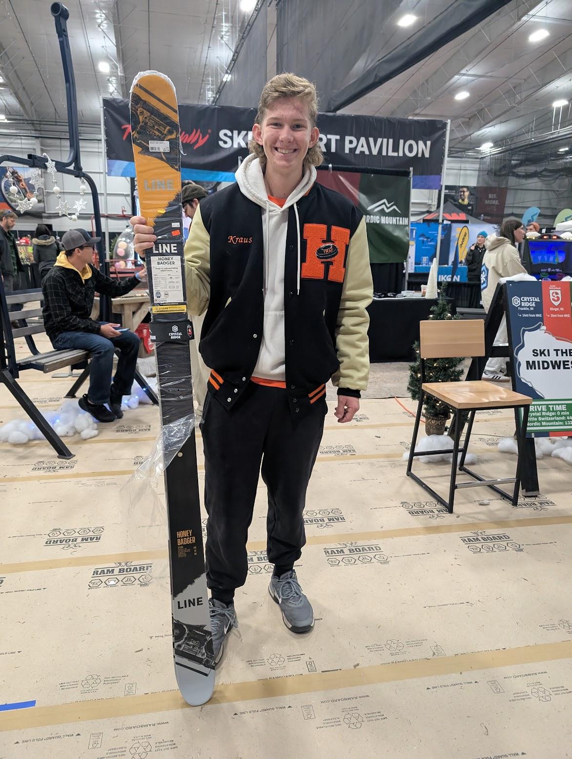 Man in black clothing stands on skis at a busy indoor trade show booth.