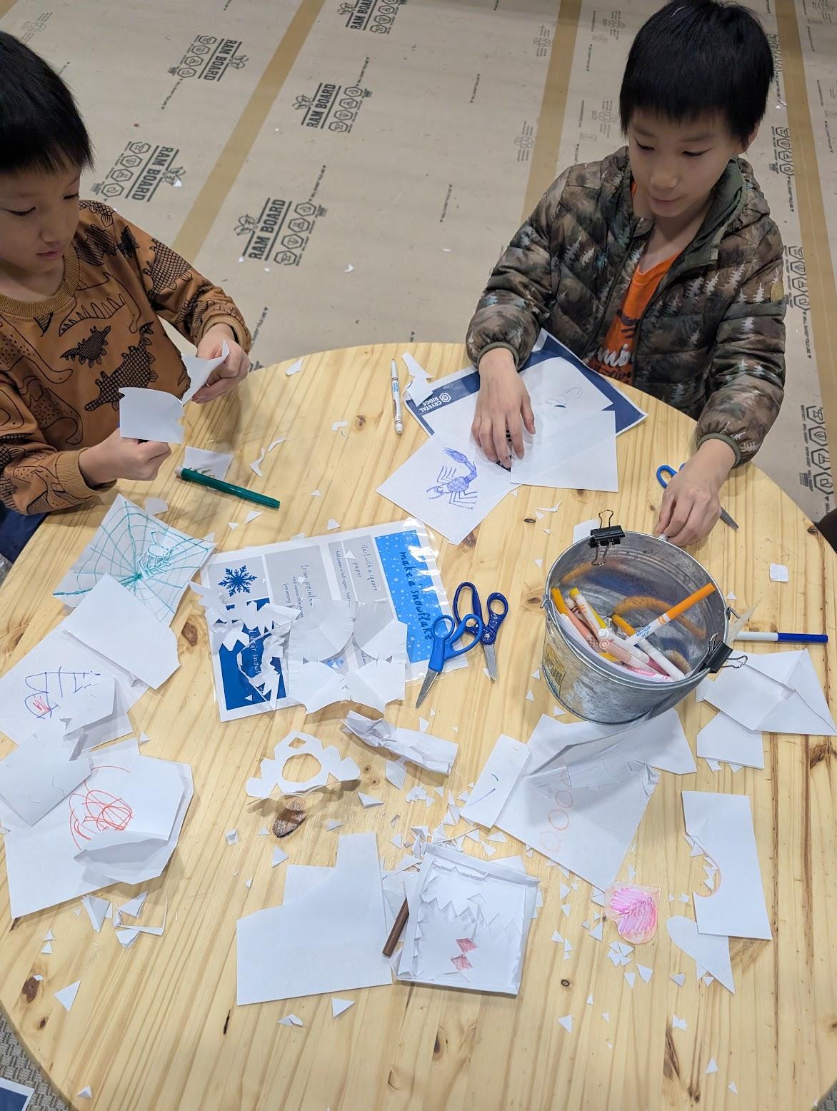 Two children crafting with paper, scissors, and markers on a wooden floor.