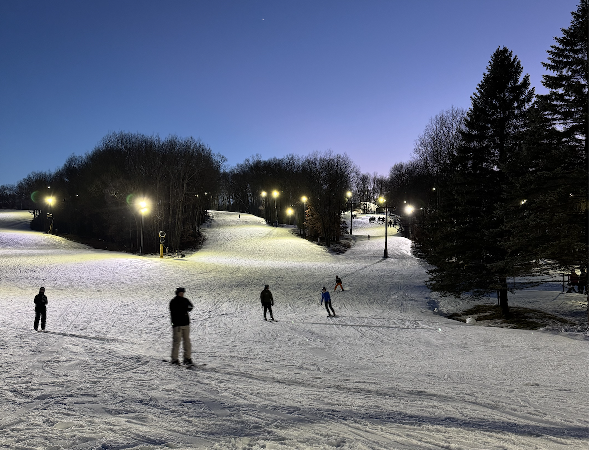 People skiing and walking on a snowy hill at dusk under streetlights.