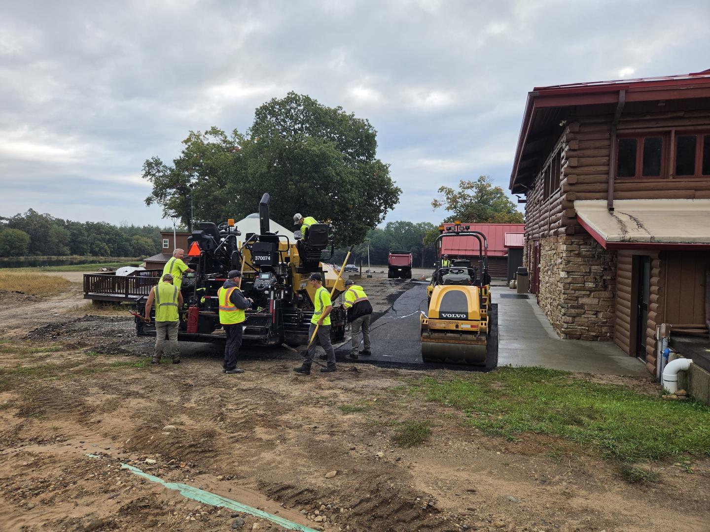Construction workers and machinery beside a wooden building under cloudy skies.