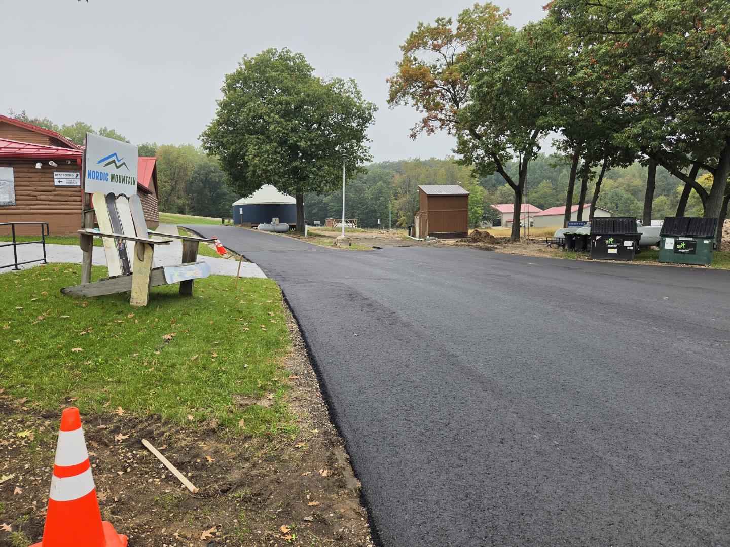 Freshly paved road beside a grassy verge with orange traffic cone and parked vehicles.