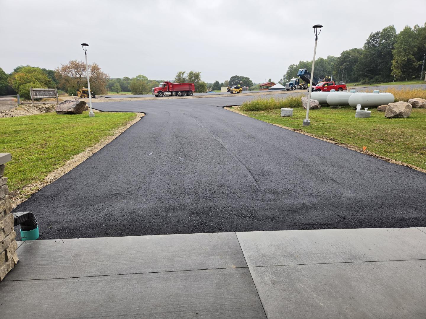 Freshly paved road leading past parked cars and grassy lots under a cloudy sky.