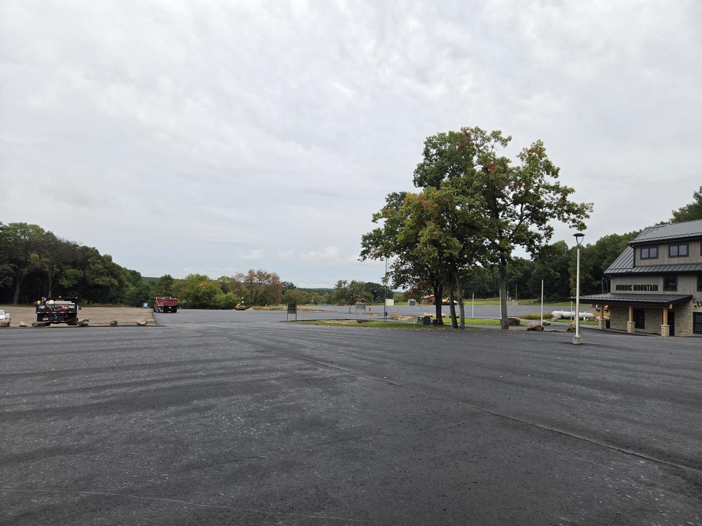 Nearly empty asphalt parking lot with trees and buildings under a cloudy sky.