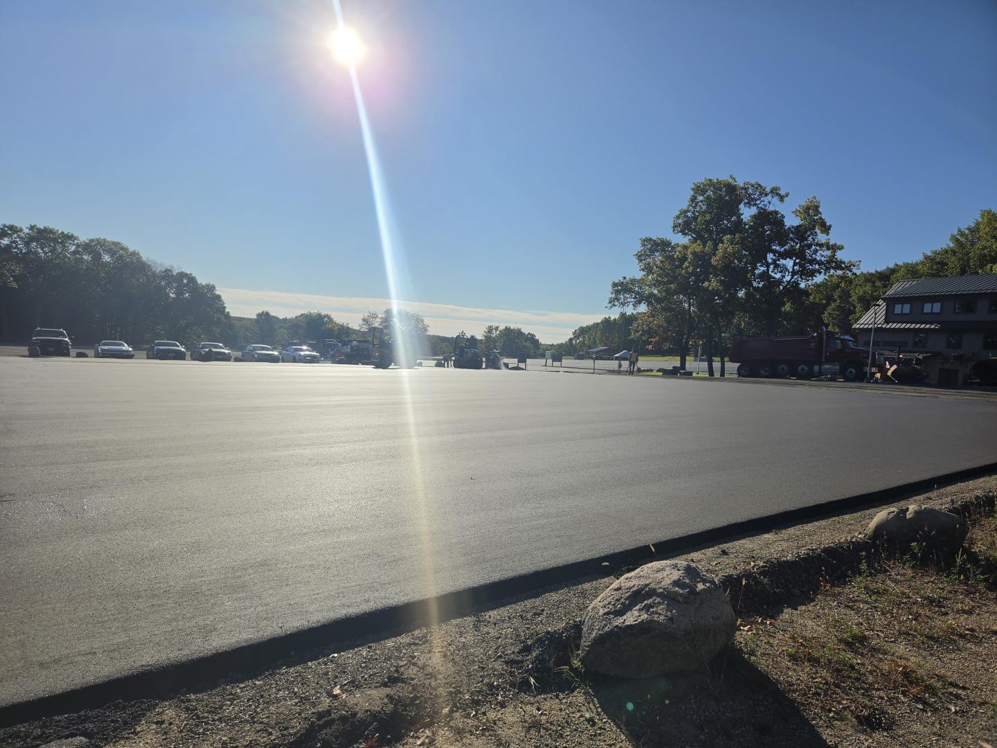 Sunlit parking lot with trees and a building in the distance.