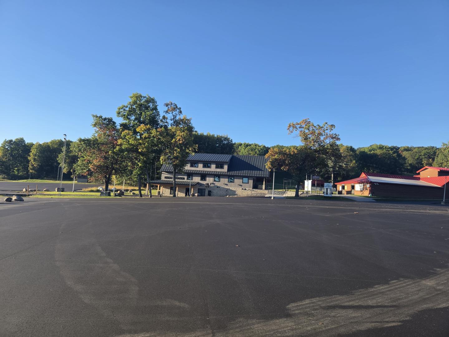 Empty asphalt parking lot in front of a low building and trees under a clear blue sky.