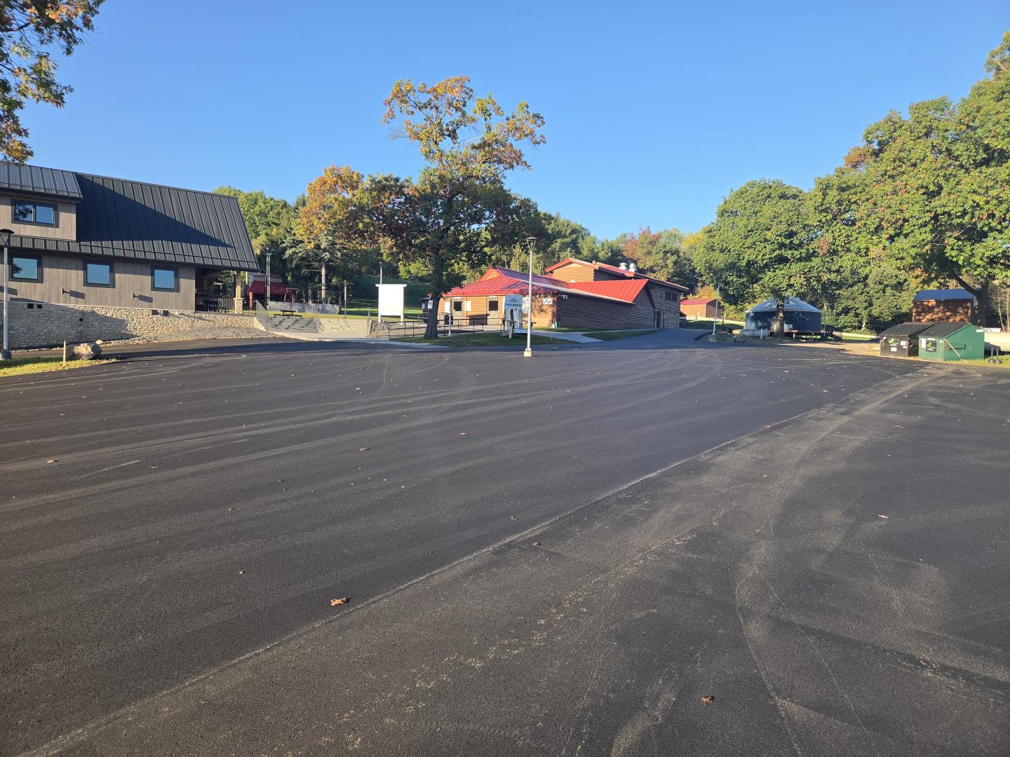 Freshly paved empty parking lot beside low buildings and leafy trees under a clear blue sky.