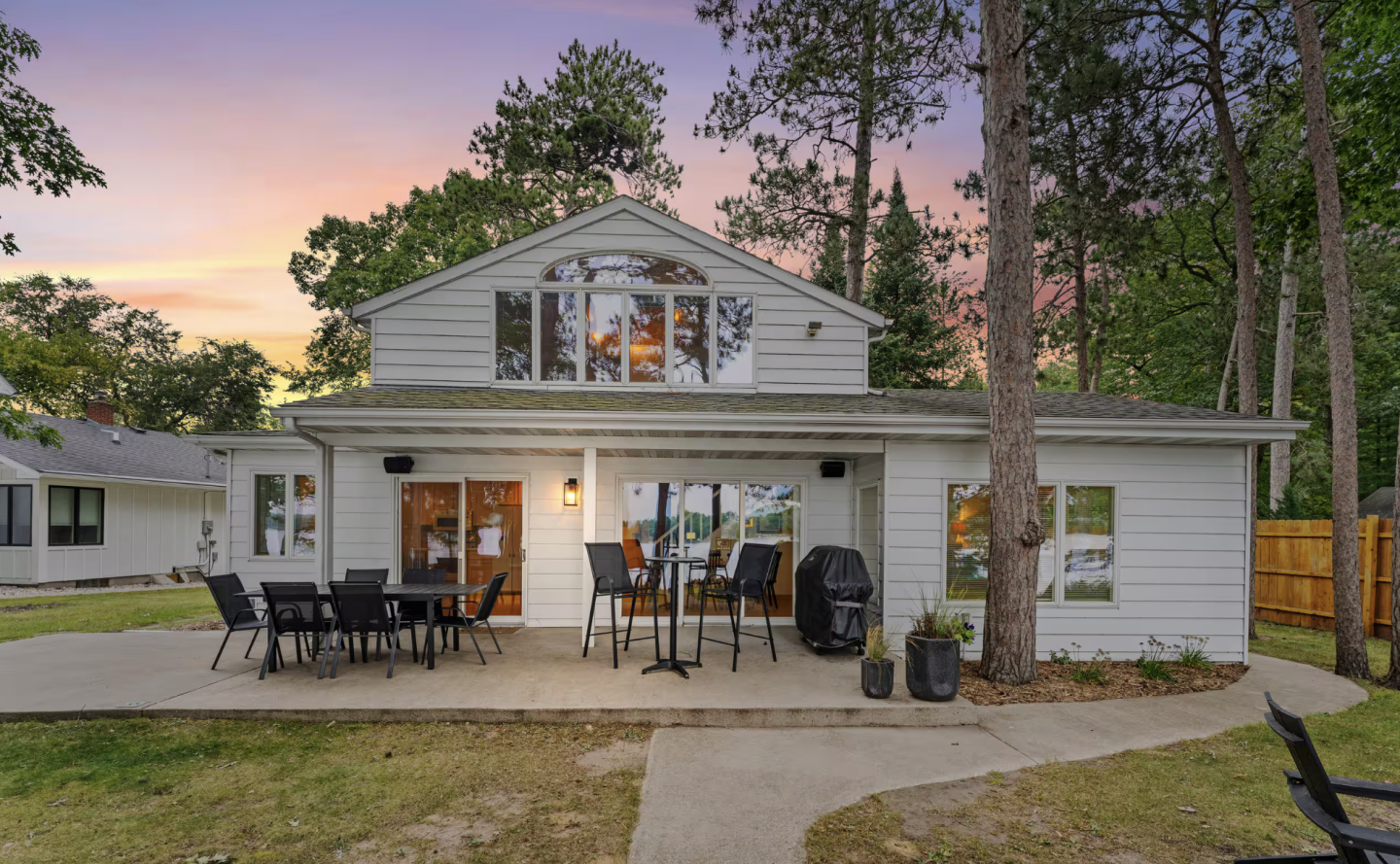 White farmhouse with a covered patio at sunset, surrounded by tall trees.